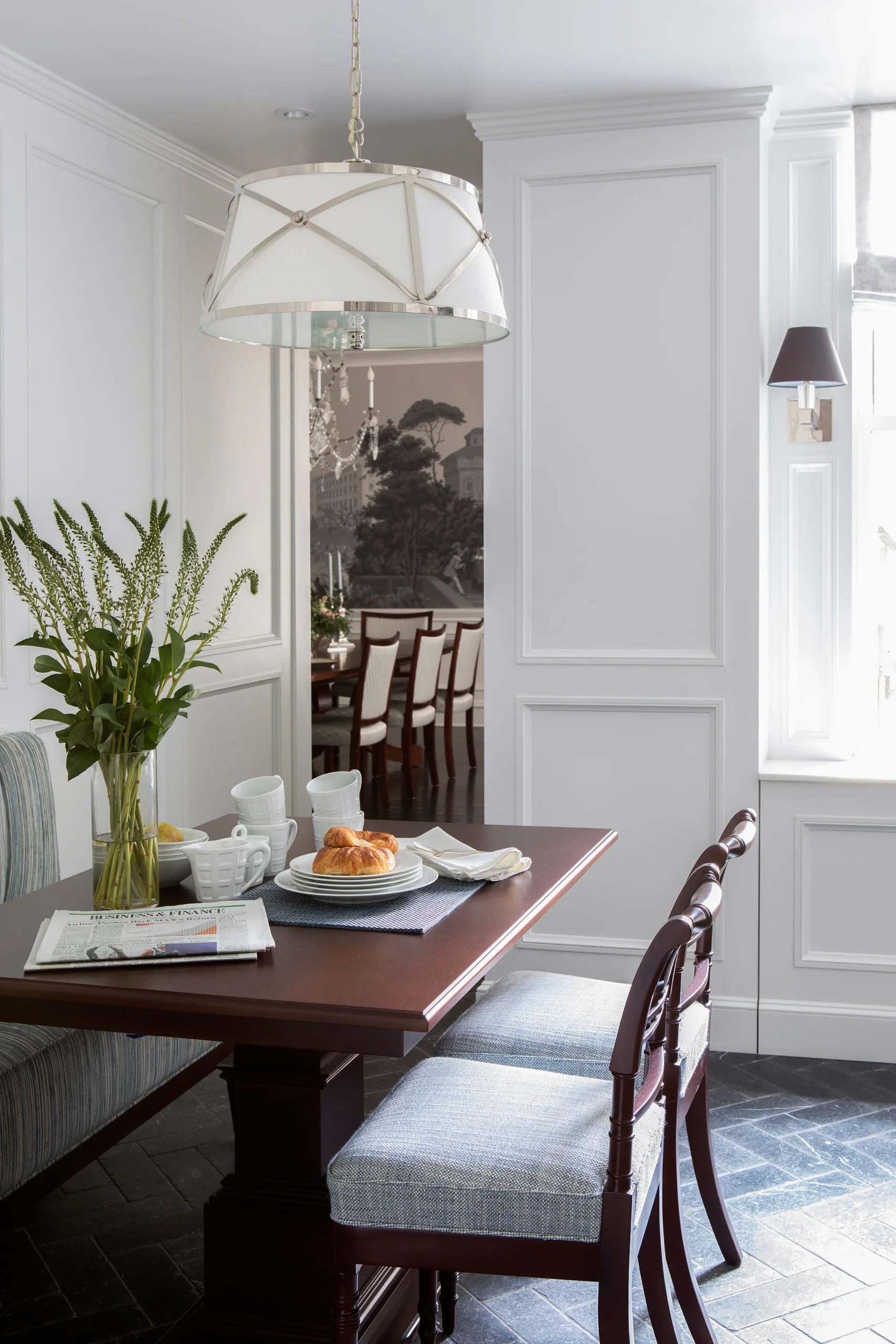 A warm and inviting dining nook featuring a dark wood table set for breakfast with croissants and a tea set. The space is framed by soft natural light, a statement pendant light fixture, and a vase of fresh greenery, creating the perfect spot for a relaxing meal.