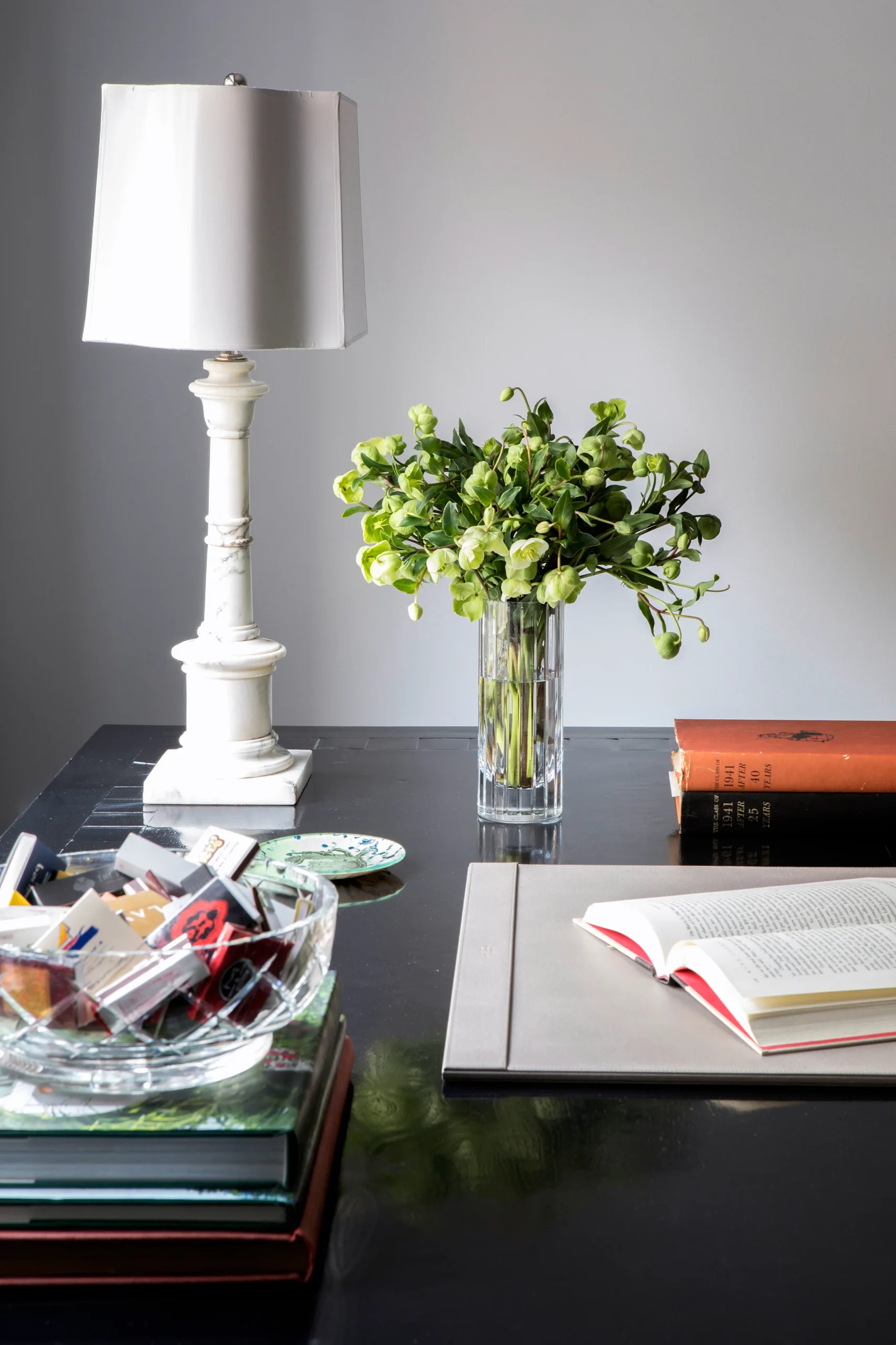 A sophisticated office setup with a sleek black desk, featuring an open book, a white lamp, and a vase of fresh green flowers. A crystal bowl filled with colorful matchboxes adds a creative touch, completing this elegant and productive workspace.