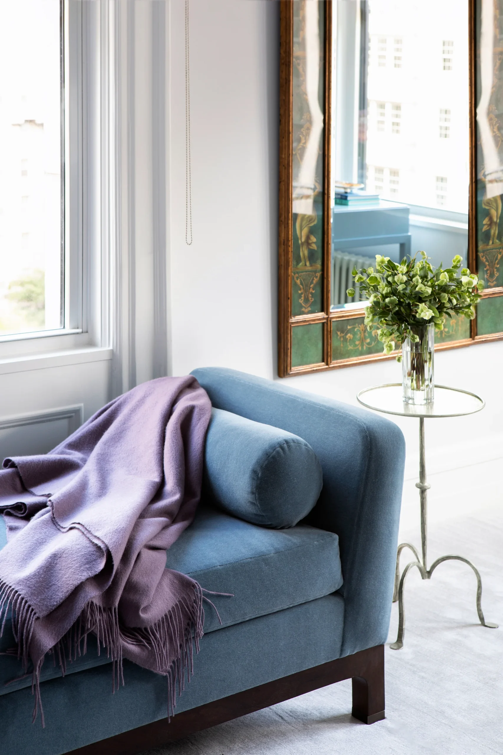 A luxurious blue chaise lounge adorned with a soft purple blanket offers a cozy retreat by the window. The decorative mirror and fresh green bouquet on a side table enhance the serene atmosphere, making this space ideal for relaxation.