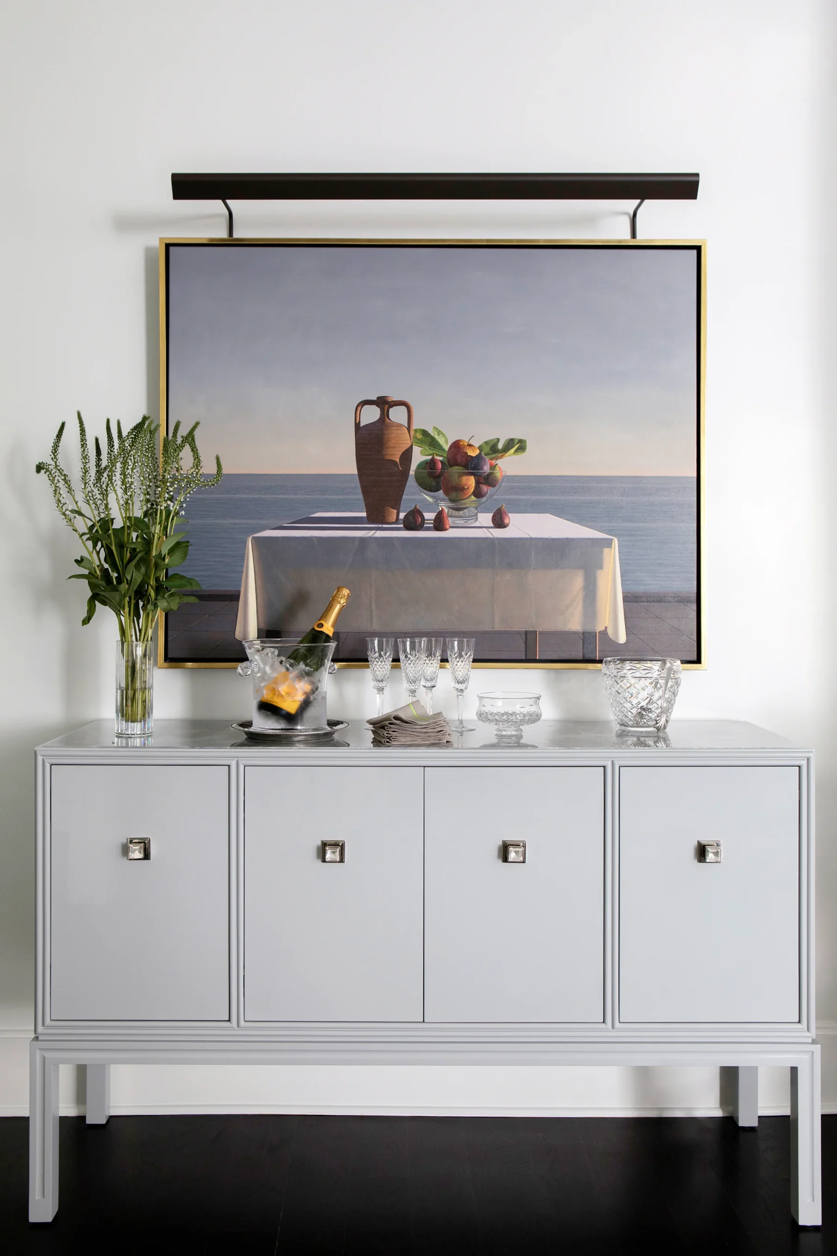 A sleek sideboard adorned with crystal glassware and a bottle of champagne on ice, set against a serene still-life painting. The minimalist design of the cabinet complements the artistic elements, creating a luxurious yet understated vignette. A vase of fresh greenery adds a touch of nature to this sophisticated arrangement.