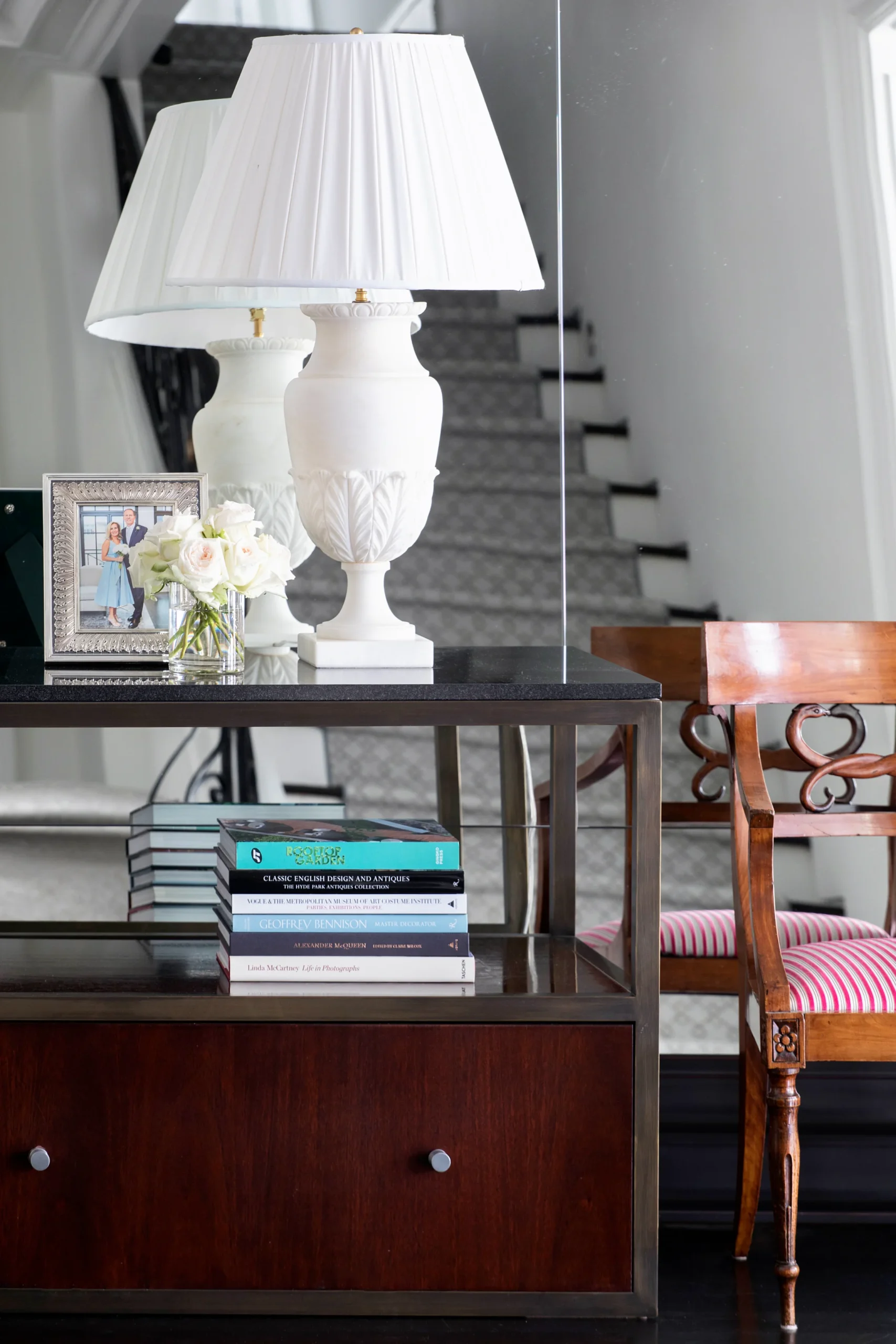 A stylish hallway console featuring a classic white lamp, a framed family photo, and a vase of fresh flowers. Below, a collection of design and photography books adds a touch of intellectual charm, while the reflection in the mirror and the nearby staircase lend depth and elegance to the scene.