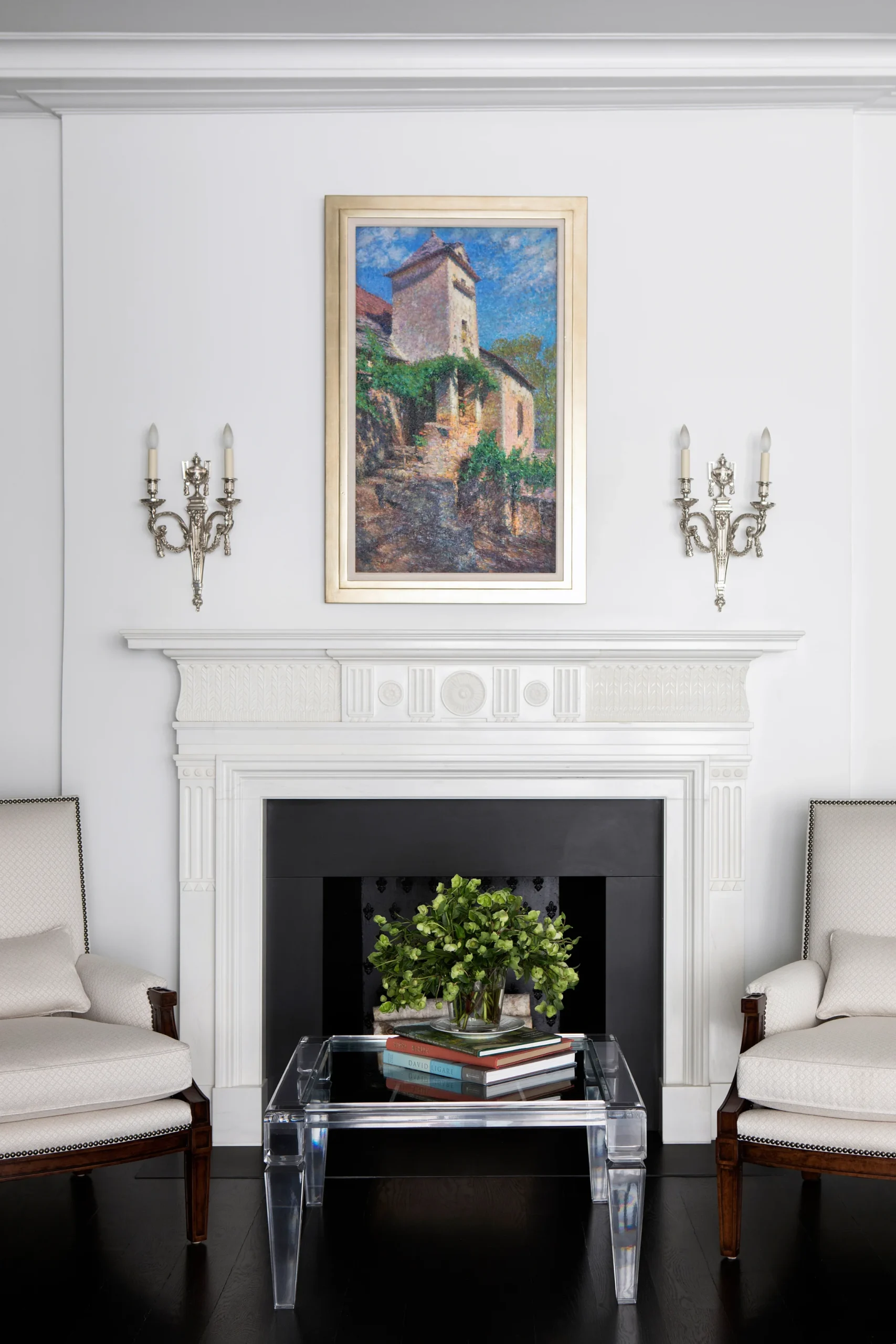 A refined living room showcasing a classic white fireplace framed by intricate molding, complemented by silver sconces and a beautiful landscape painting. The clear acrylic coffee table with a floral arrangement adds a modern touch to the space, balancing the traditional design elements with contemporary style.