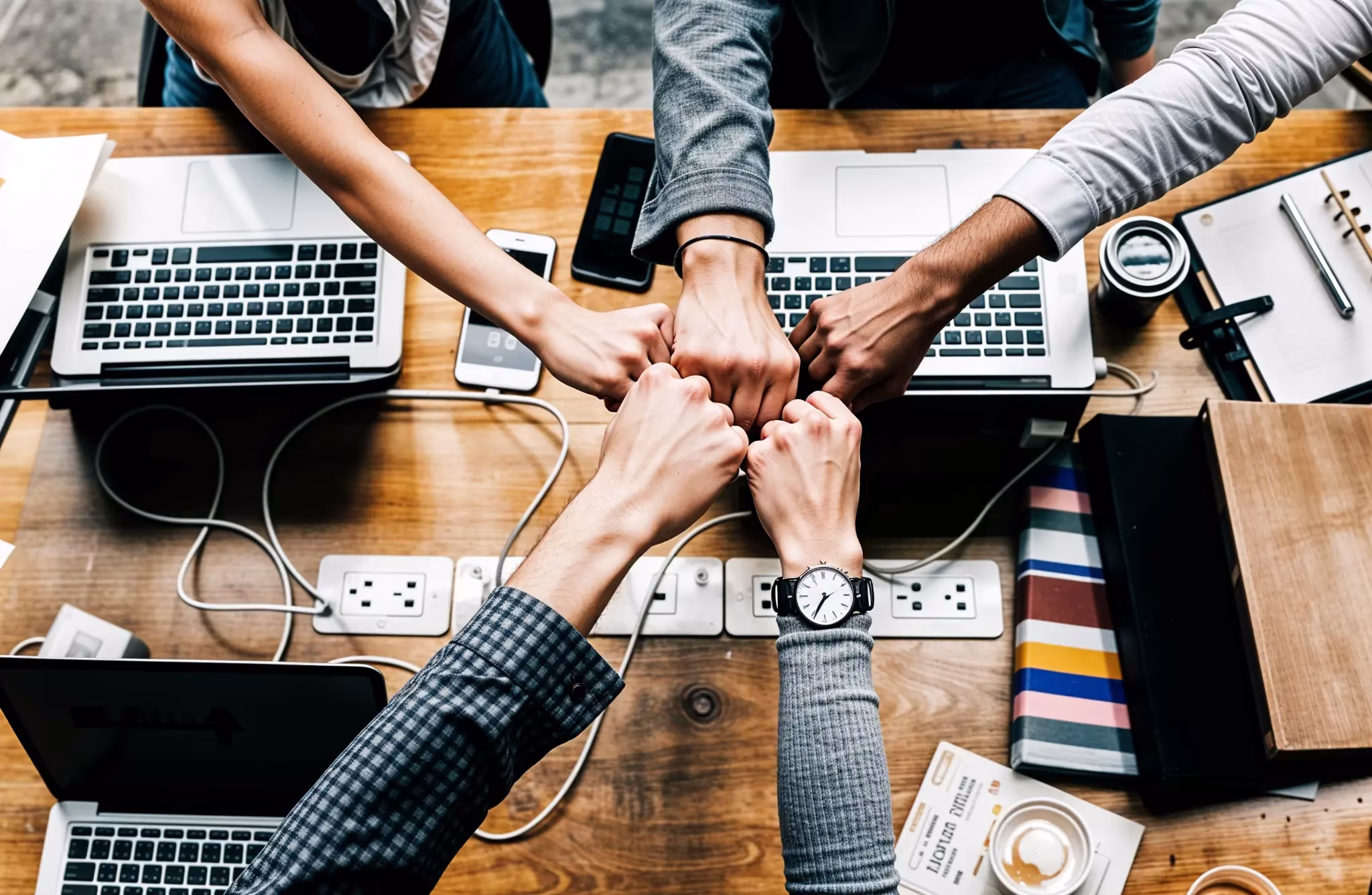 Team of professionals collaborating with a fist bump around a wooden desk filled with laptops and documents.