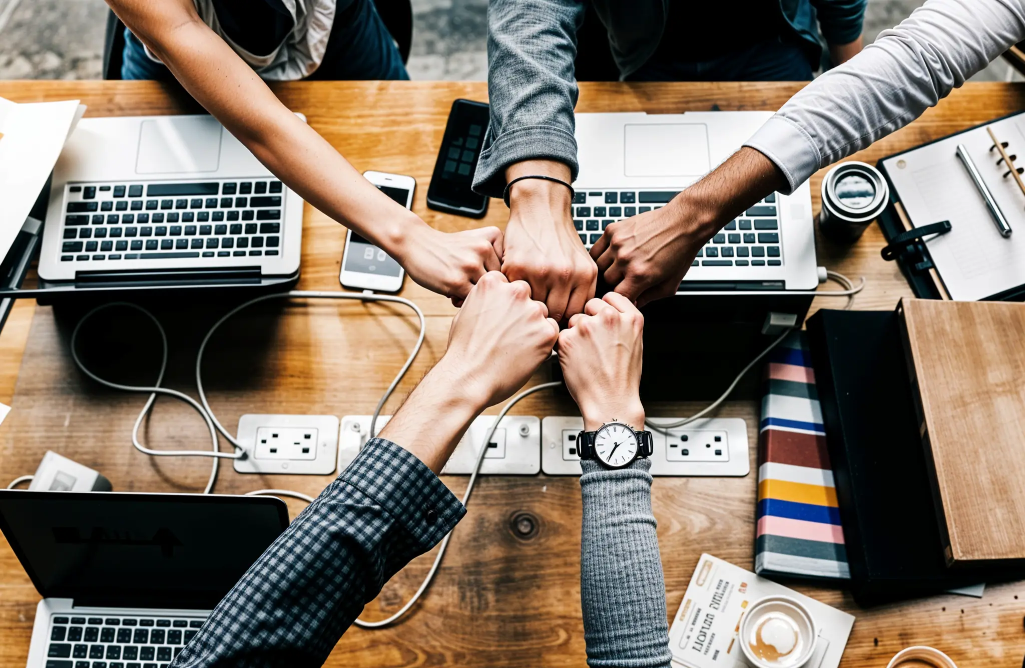 Team of professionals collaborating with a fist bump around a wooden desk filled with laptops and documents.